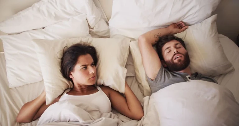 A imagem mostra um casal deitado na cama, enquanto o homem dorme e ronca a esposa está acordada irritada com o ronco.